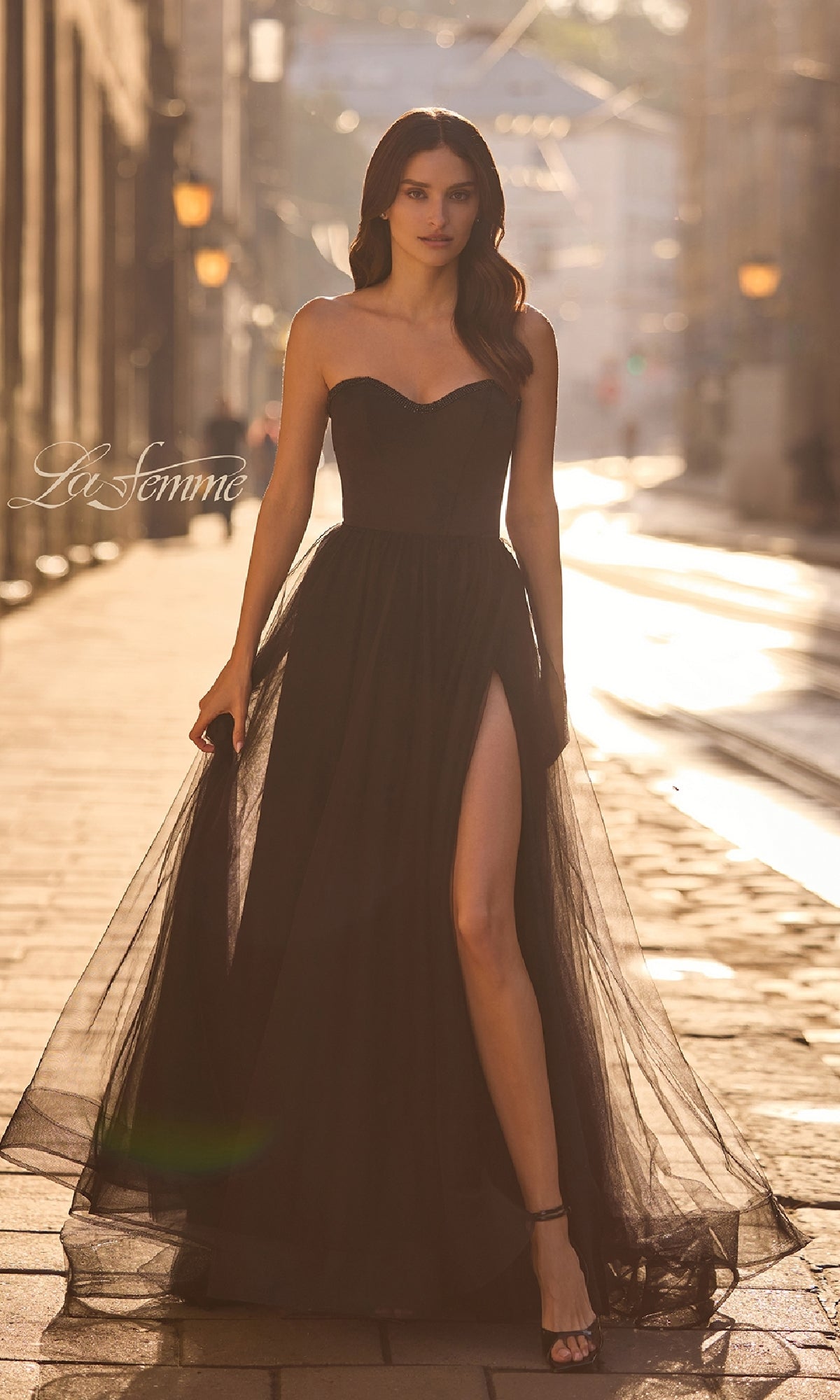 A woman wears the La Femme 32731 Strapless Long Prom Ball Gown with a sweetheart neckline, sheer skirt, and thigh-high slit, paired with black heels on a sunlit street.